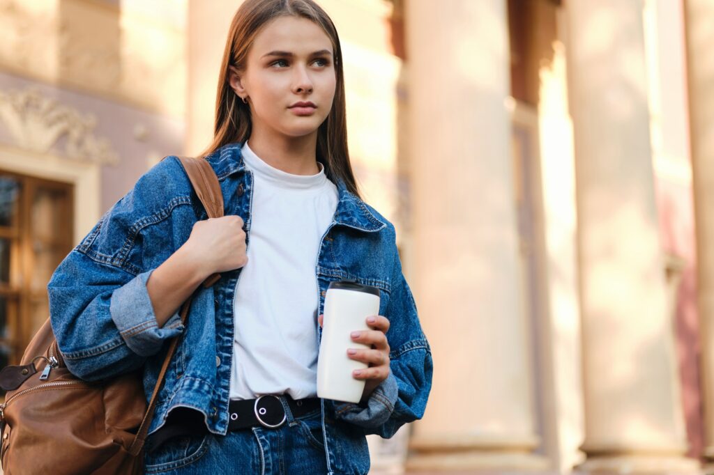 Attractive stylish student girl in denim jacket with backpack thoughtfully looking away outdoor