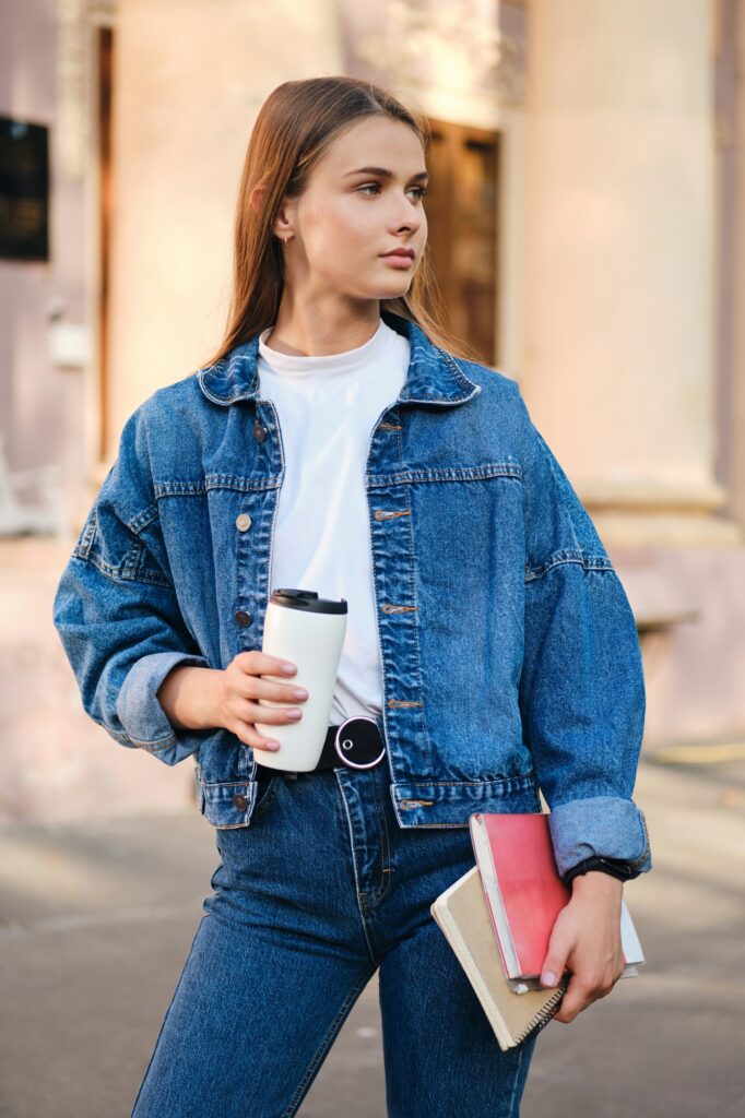 Attractive stylish student girl in denim jacket with coffee thoughtfully looking away outdoor